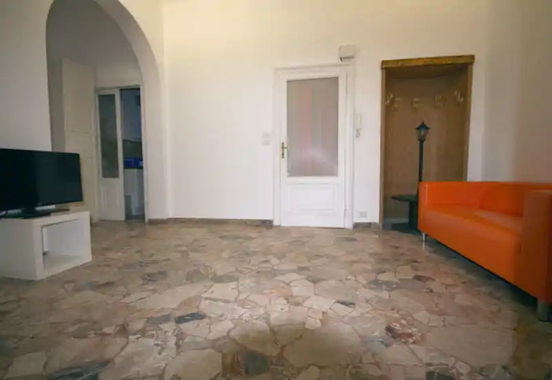 Ampio soggiorno con pavimento in pietra chiara, divano arancione e angolo TV; layout semplice e arioso con arco d'ingresso e nicchia appendiabiti.