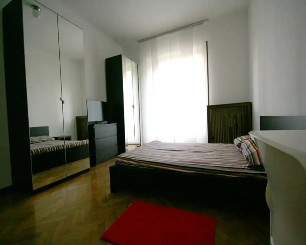 Dormitorio luminoso y sencillo con cama doble, armario con puertas espejadas y suelo de parquet. Gran ventana con cortina traslúcida que aporta luz; ropa de cama a rayas y una pequeña alfombra añaden color.