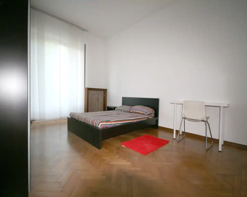 Dormitorio minimalista con cama baja de estructura oscura, ropa de cama a rayas, una pequeña alfombra roja y escritorio junto a una ventana luminosa. Suelo de parquet y paredes neutras que crean un espacio tranquilo y aireado, perfecto para anuncios.