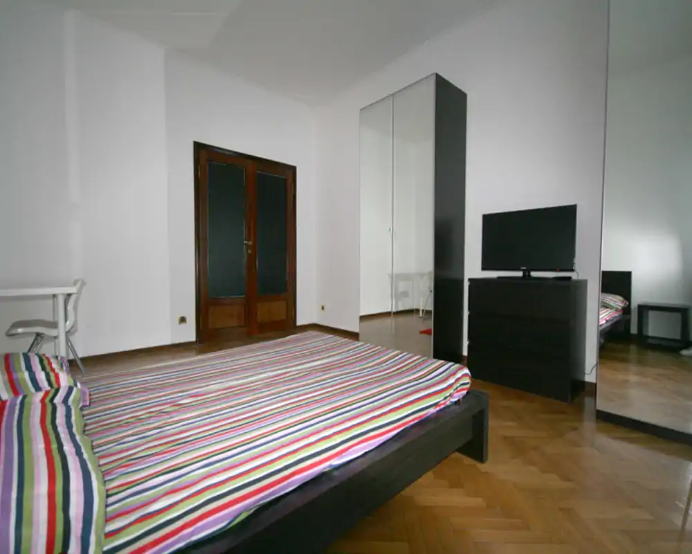 Dormitorio amplio y sencillo con cama baja de matrimonio con ropa de cama de rayas coloridas, armario con espejo y cómoda con televisor. Suelo de parquet cálido y paredes neutras crean un ambiente luminoso y ordenado.