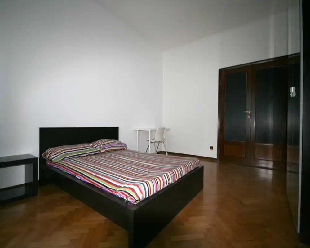 Dormitorio minimalista con cama plataforma negra y edredón a rayas multicolor, suelo de parquet y paredes blancas: espacio luminoso y despejado, perfecto para anuncios de alquiler.