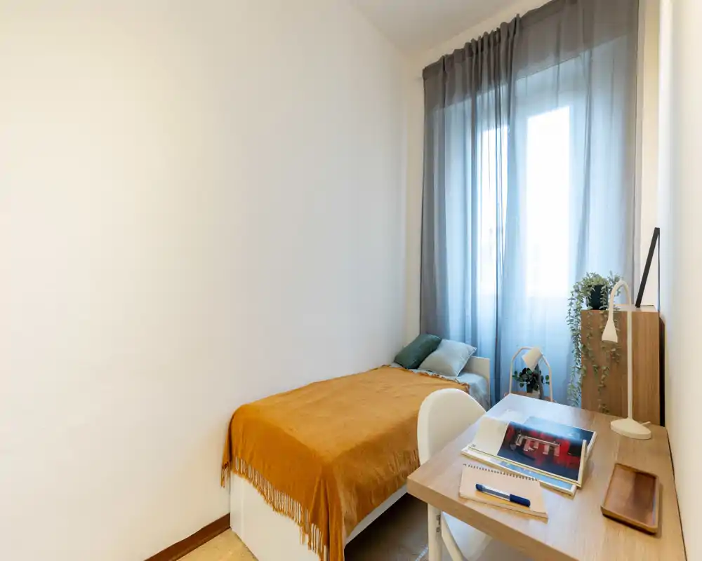 Compact, well-lit bedroom with a single bed dressed in a warm orange throw, a desk by the window and soft natural light filtered through sheer curtains.