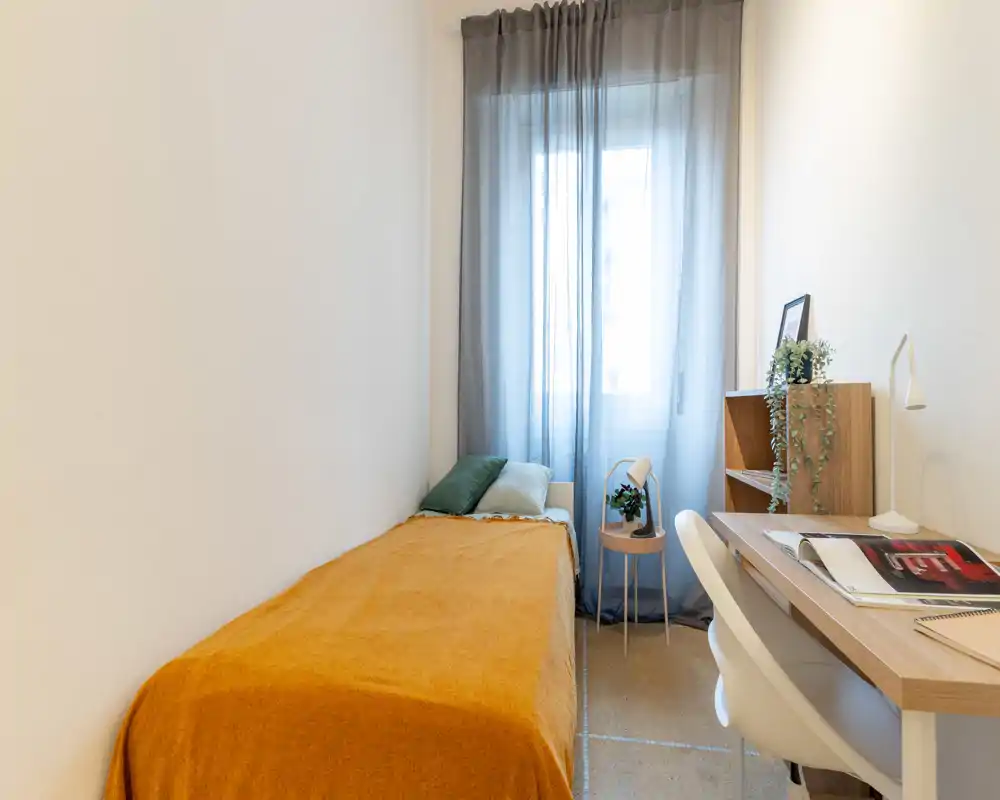 Compact, well-lit single bedroom with a mustard throw, pillows and a small desk beside the window; neat, cozy layout ideal for a student or short-term stay.