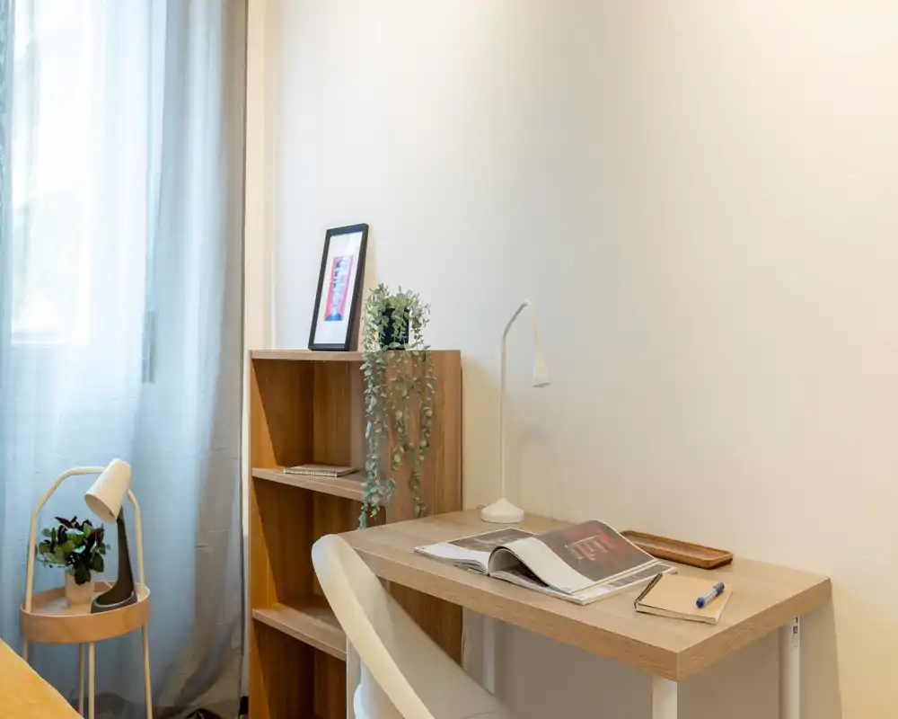 Bright, minimal bedroom corner with a wooden desk, shelving and soft natural light; tasteful plants and a reading lamp create a calm workspace.