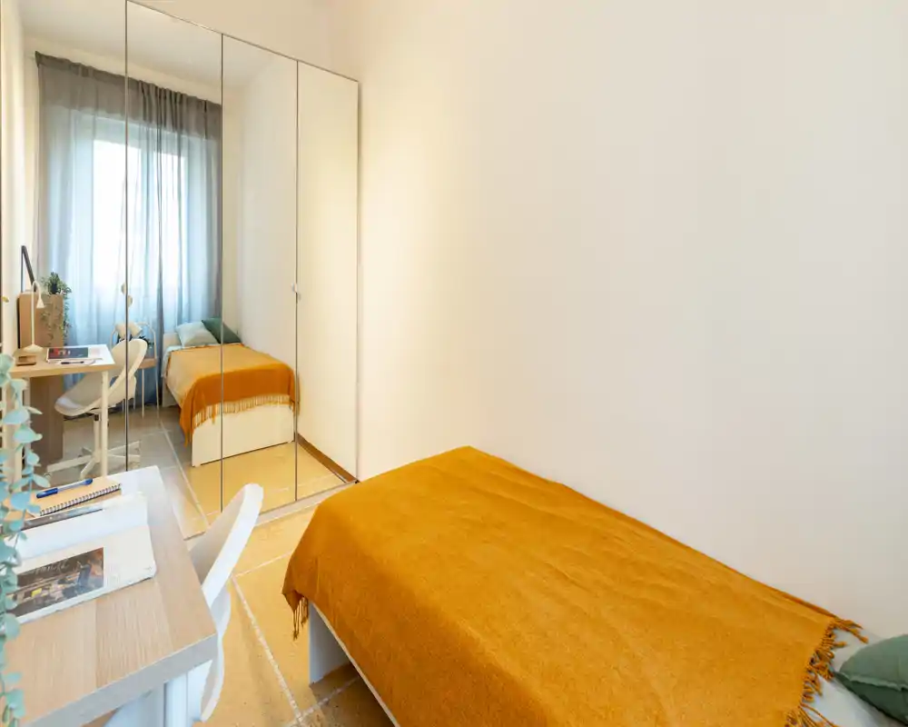 Small, well-lit single bedroom with a mustard throw, mirrored wardrobe and a compact desk by the window creating a bright, cozy workspace.