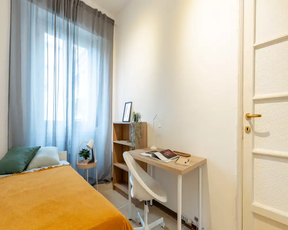 Well-lit compact bedroom with a single bed, mustard throw and green cushions, a small desk and shelving — ideal for a student or single occupant.