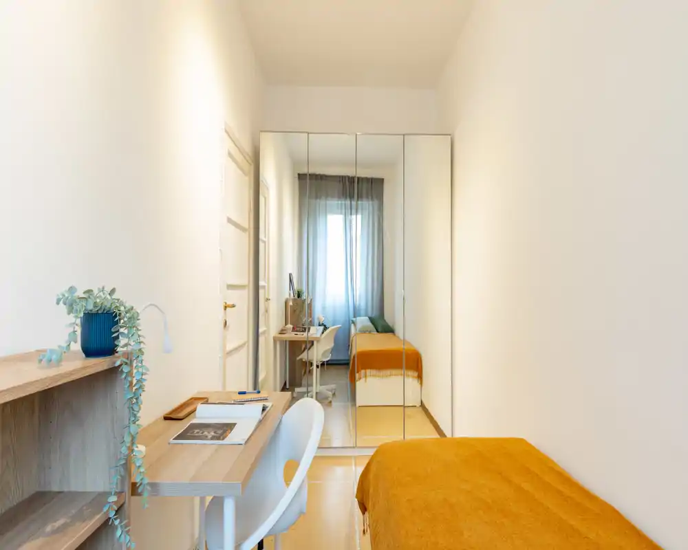 Compact, well-staged bedroom with a single bed dressed in a warm orange throw, a desk and chair for study, and mirrored wardrobes that add depth and light.