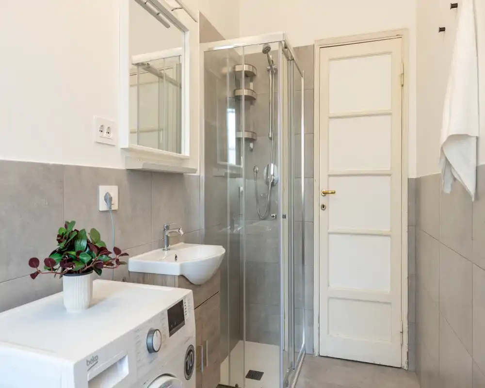 Modern bathroom with a glass shower enclosure, wall-mounted sink, washing machine and neutral grey tiles — bright and clean.