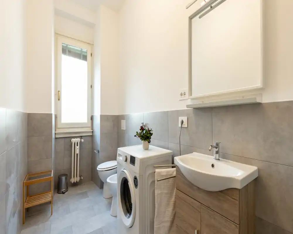 Bright, modern bathroom with a sink, mirror cabinet, toilet and a front-loading washing machine; neutral gray tiles and wooden vanity give a clean, contemporary look.