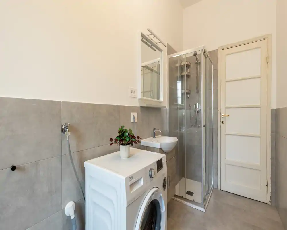 Bright, modern bathroom with a glass shower cabin, small sink and a washing machine — clean neutral tiles and minimal decor.