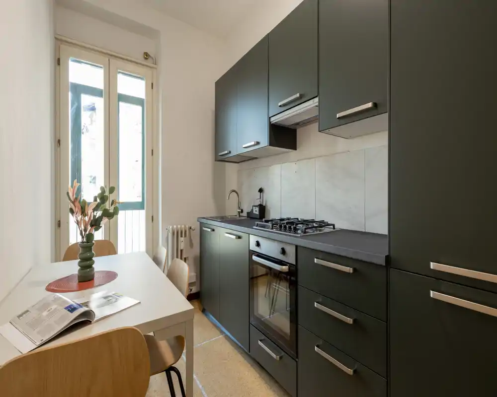 Modern compact kitchen with dark matte cabinets, built-in oven and gas hob, and a small dining table by a tall window providing natural light.