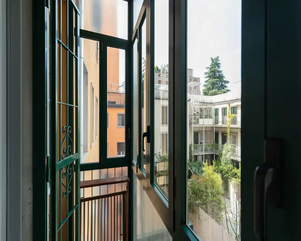 Small enclosed balcony with green metal framed windows opening onto a courtyard; views of neighboring buildings and greenery create a bright, private outdoor nook.