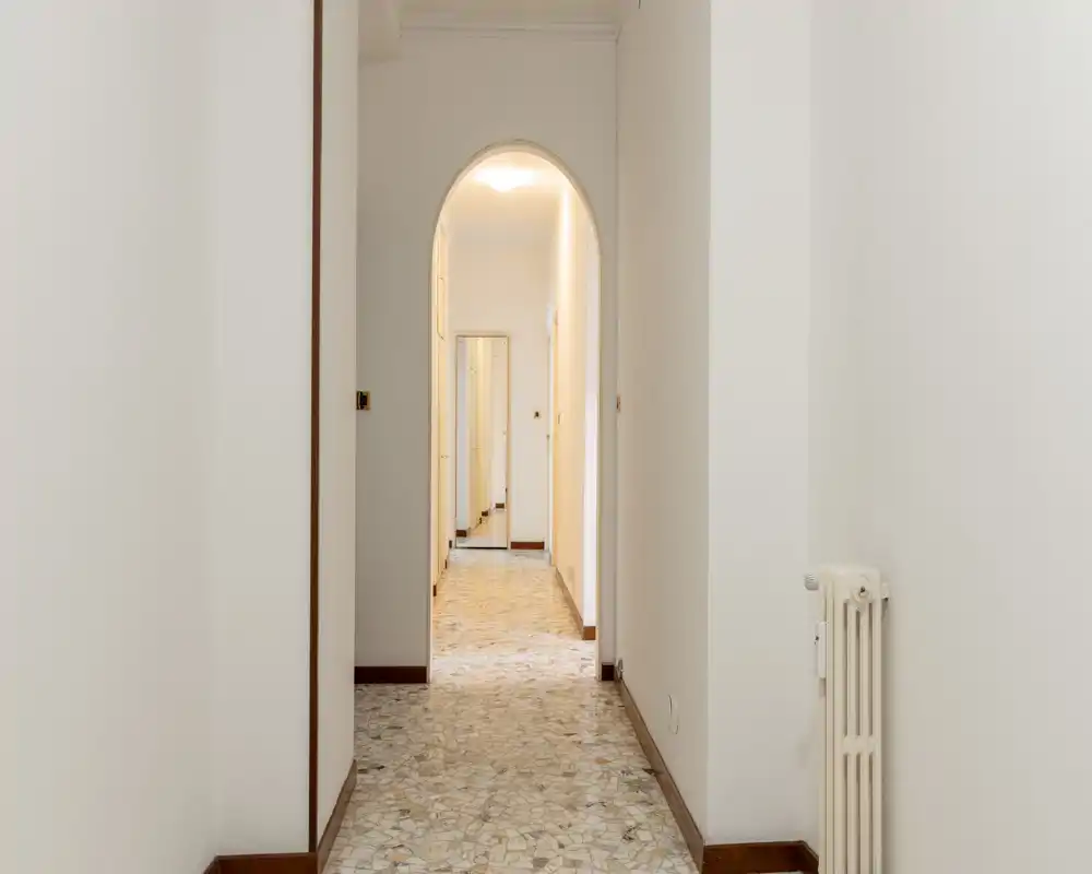 Long, bright entrance corridor with terrazzo floor, white walls and an elegant arched doorway leading to inner rooms — clean and spacious transitional space.