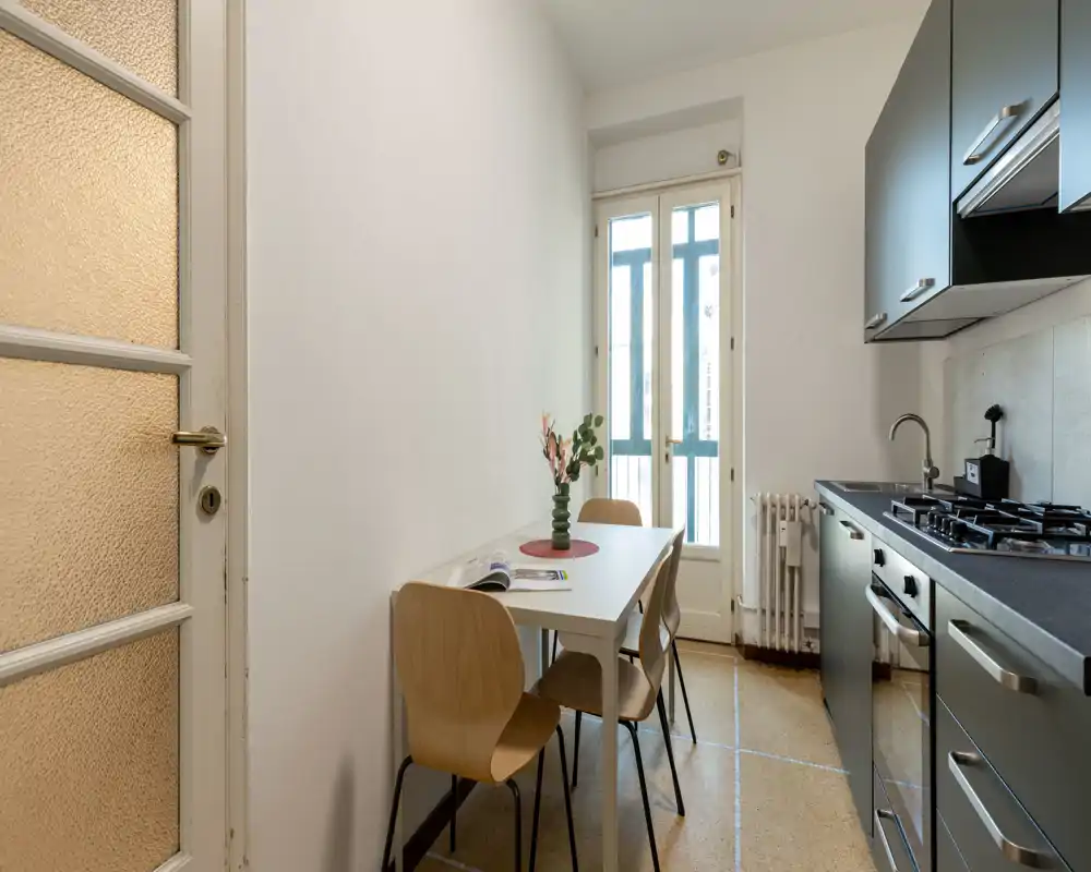 Compact modern kitchen with a small dining table for two, natural light from the glazed door, and dark cabinetry with built-in appliances — ideal for a cozy city apartment.