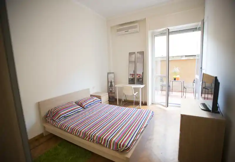 Chambre lumineuse et simplement meublée avec un lit double, table de chevet, petit bureau et accès direct à un balcon apportant la lumière naturelle.