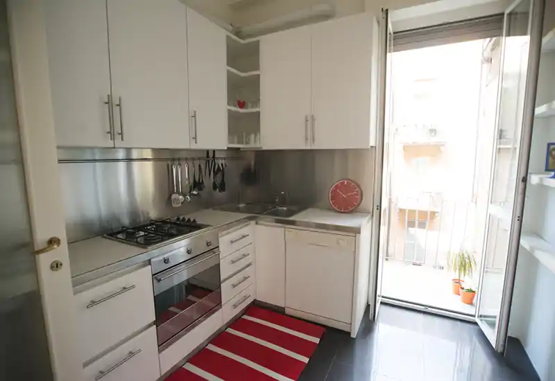 Cuisine moderne et lumineuse avec meubles blancs, crédence en inox, four encastré et plaque à gaz. La lumière naturelle entre par la porte-fenêtre du balcon ; un tapis rayé rouge et des accessoires apportent de la chaleur.