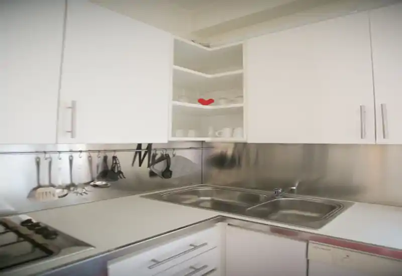 Cuisine moderne et compacte avec évier et crédence en inox, meubles blancs, ustensiles suspendus et petit cœur décoratif sur une étagère ouverte — espace lumineux et soigné, parfait pour une annonce.