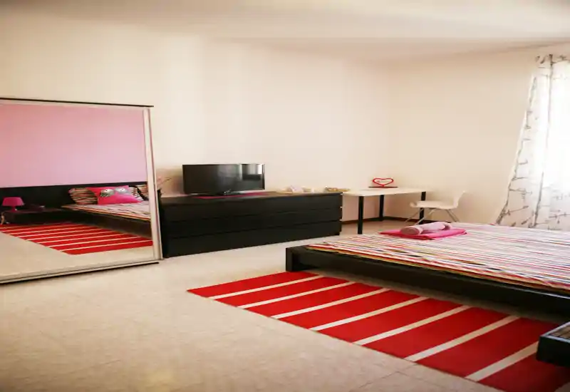 Camera da letto luminosa e moderna con letto basso a piattaforma, biancheria a righe e tappeto rosso coordinato; grande armadio con specchio e comò per spazio e profondità.