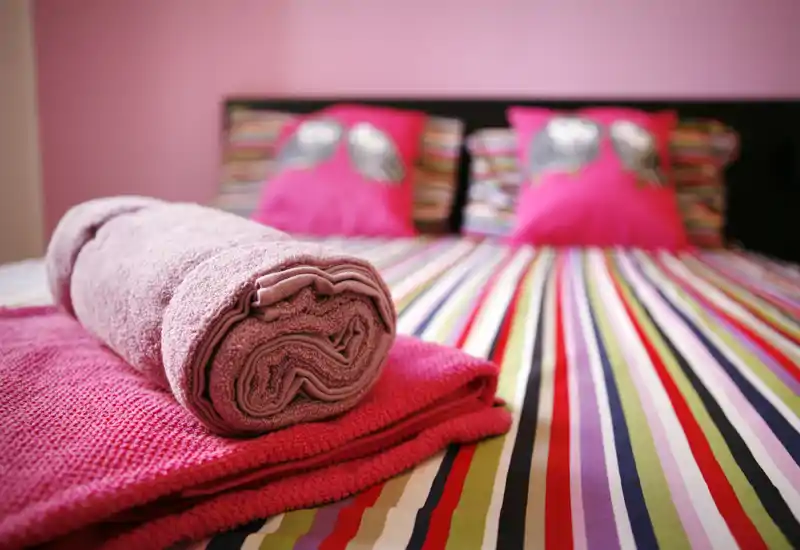 Camera da letto luminosa e accogliente con copriletto a righe colorate, cuscini rosa decorativi e asciugamani piegati in primo piano — perfetta per un annuncio di affitto.