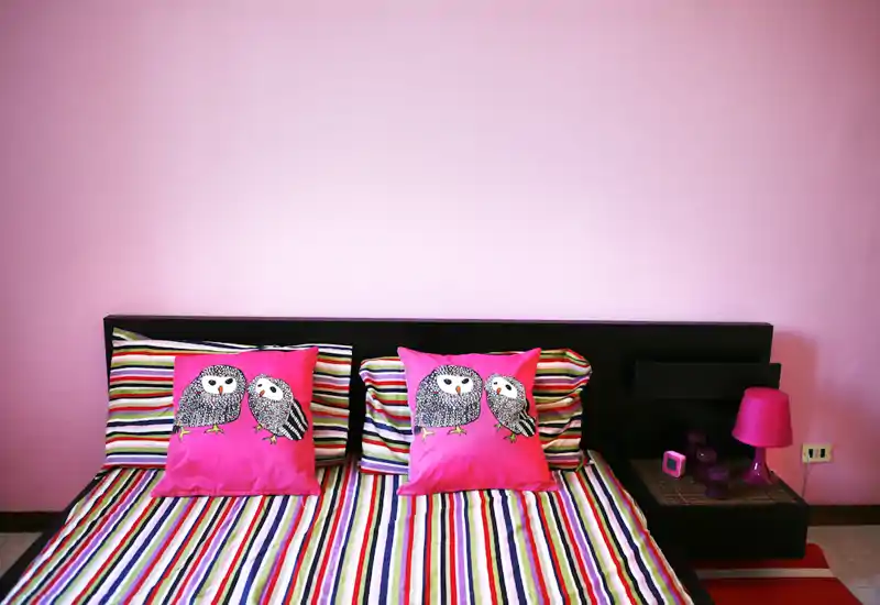 Camera da letto luminosa e vivace con copriletto a righe e due cuscini decorativi con gufi; testiera nera e comodino coordinato con lampada rosa per un ambiente curato e giocoso.
