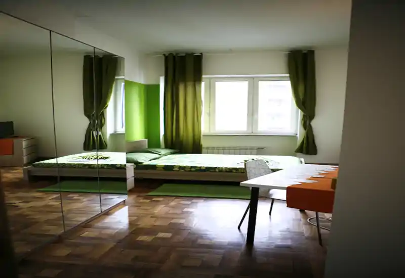Dormitorio moderno y luminoso con cama baja tipo plataforma, acentos verdes y un gran armario con espejo que amplía el espacio. La luz natural, las alfombras y las cortinas coordinadas crean un ambiente fresco y acogedor.