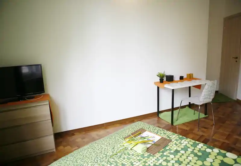 Dormitorio luminoso y sencillo con colcha verde estampada, pequeño escritorio y silla, cómoda con televisión y suelo de parquet. La habitación está ordenada con una planta y detalles verdes que aportan un ambiente fresco y acogedor.