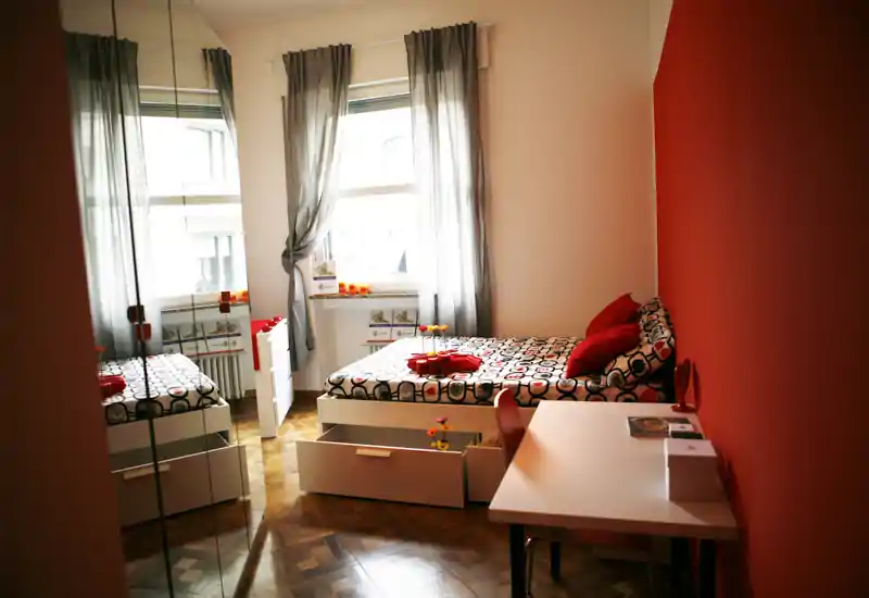 Dormitorio luminoso y con estilo, con cama individual estampada, pared roja de acento y escritorio ordenado. La luz natural y los accesorios crean un ambiente acogedor.