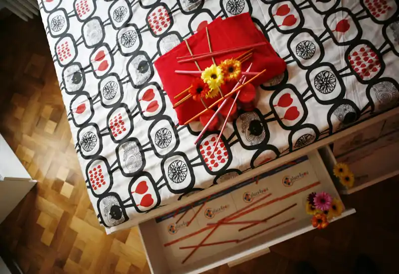 Vista desde arriba de una cama bien arreglada con ropa de cama estampada, flores decorativas y una manta roja doblada que crean una composición acogedora y colorida.