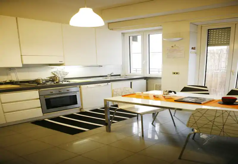 Cocina moderna y luminosa con horno empotrado, lavavajillas y mesa de comedor. Muebles blancos, suelo de baldosas y una gran ventana que aporta luz natural — zona de cocina y comedor funcional y bien presentada.