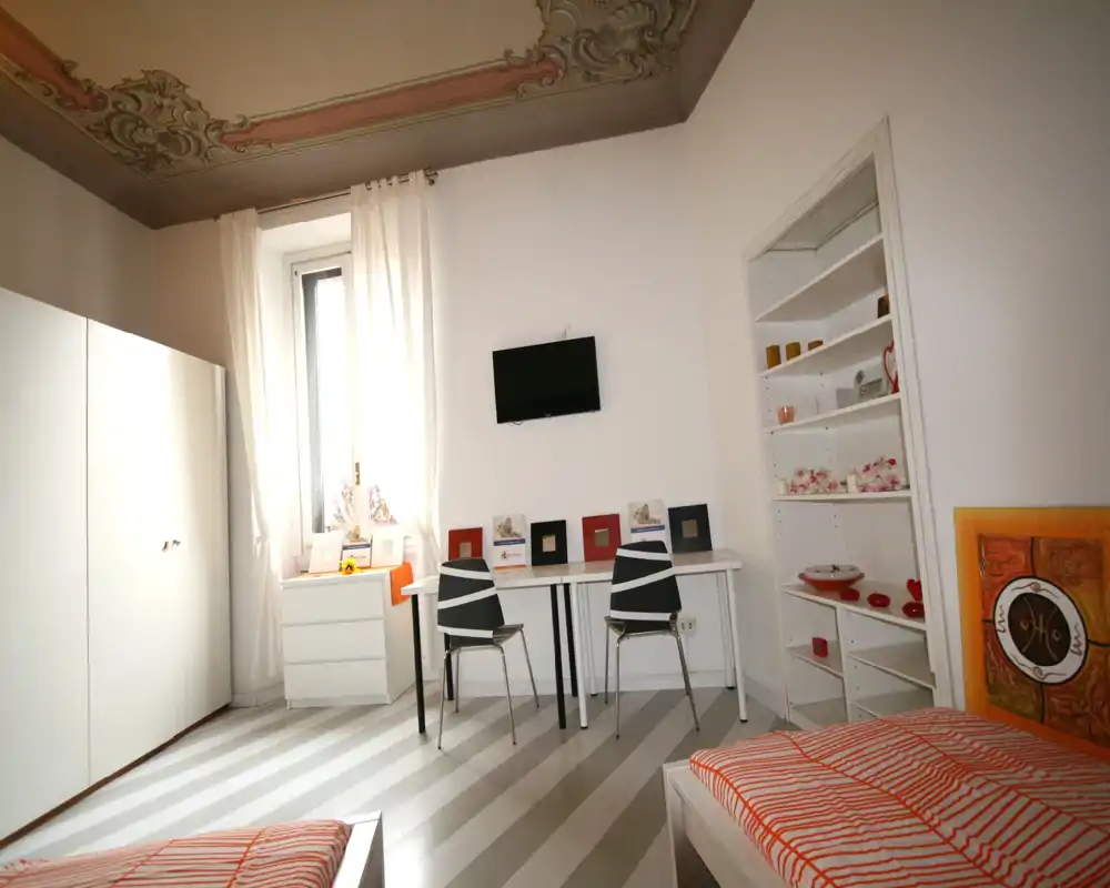 Bright twin-bedroom with two single beds, a wall-mounted TV and a desk area; decorative ceiling and striped floor add character.