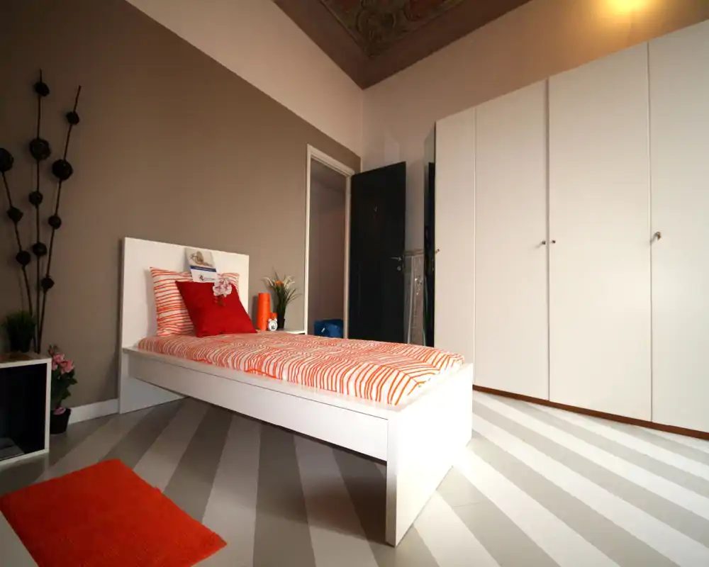 Bright, modern single bedroom with a white platform bed dressed in striped orange bedding, coordinated cushions and a large white wardrobe; clean lines and warm decor create an inviting space ideal for a guest or student.