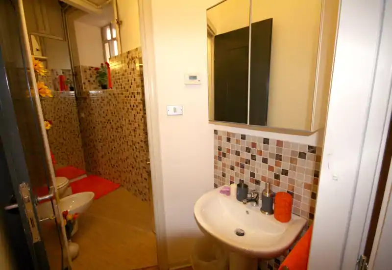 Bagno compatto e ben illuminato con piastrelle a mosaico, lavabo a colonna, specchio con mobiletto e bidet/wc visibili; illuminazione calda e dettagli arancioni conferiscono un tocco accogliente.