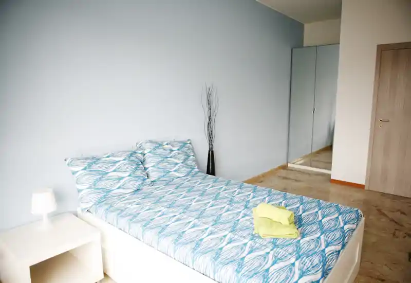 Bright, neatly staged bedroom with a double bed dressed in blue patterned linens, a bedside table with lamp, and a mirrored wardrobe reflecting the room for added depth.