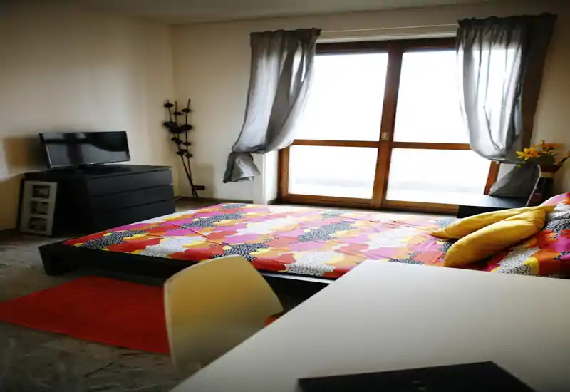 Camera da letto luminosa e ben arredata con un letto basso a piattaforma, biancheria colorata, cuscini decorativi e una finestra che illumina la stanza.