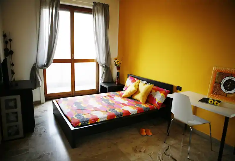 Camera da letto luminosa e moderna con letto basso nero, biancheria colorata e parete accentata gialla. Ampia finestra con tende leggere offre molta luce naturale e una scrivania crea un angolo funzionale.