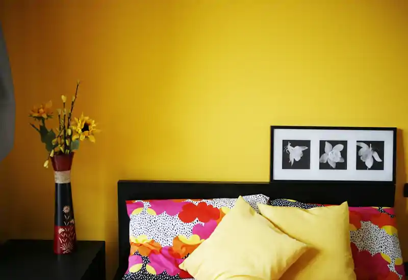 Angolo di camera luminoso e colorato con testiera decorata, cuscini gialli e un vaso di fiori coordinato su una parete gialla vivace.