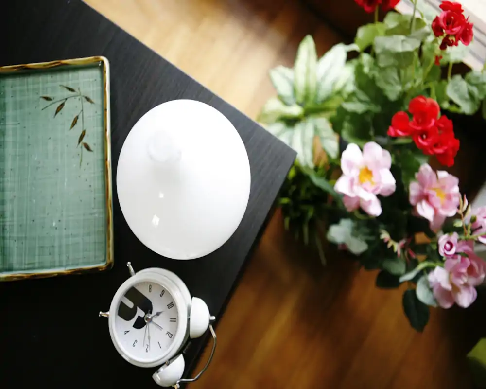 Primo piano di un comodino con lampada, sveglia e fiori decorativi su pavimento in legno; luminoso, pulito e invitante.