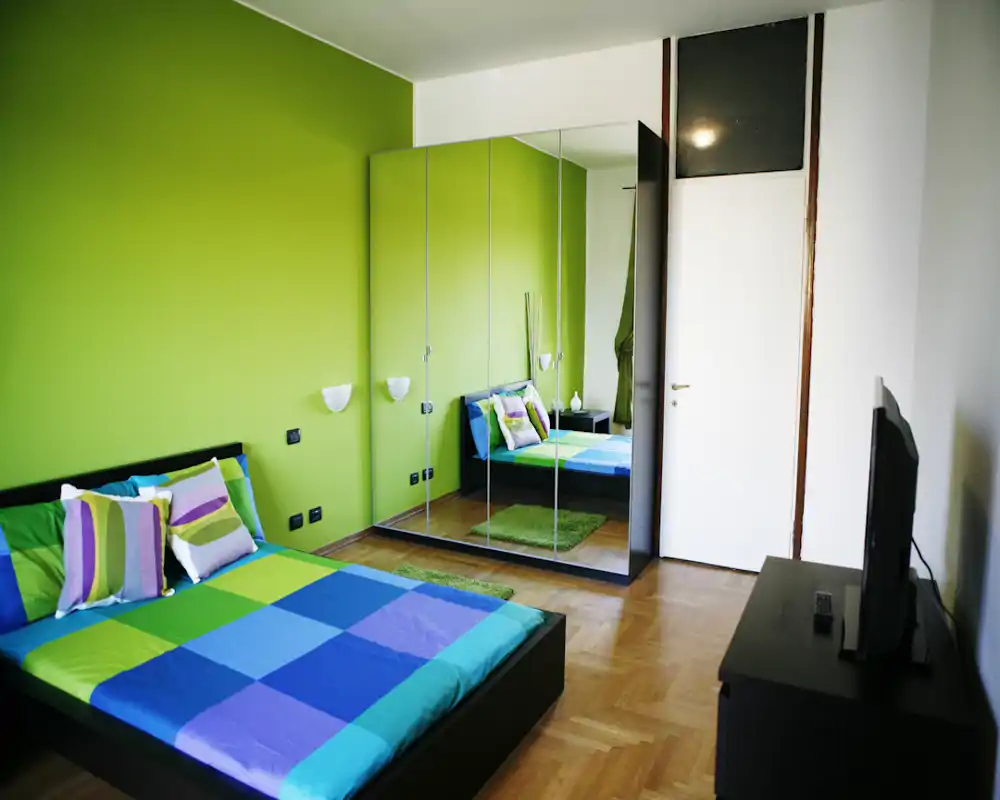 Camera da letto moderna e luminosa con letto basso e biancheria colorata blu-verde. L'armadio con ante a specchio amplia la stanza e i cuscini coordinati e il tappeto creano un'atmosfera fresca e accogliente.