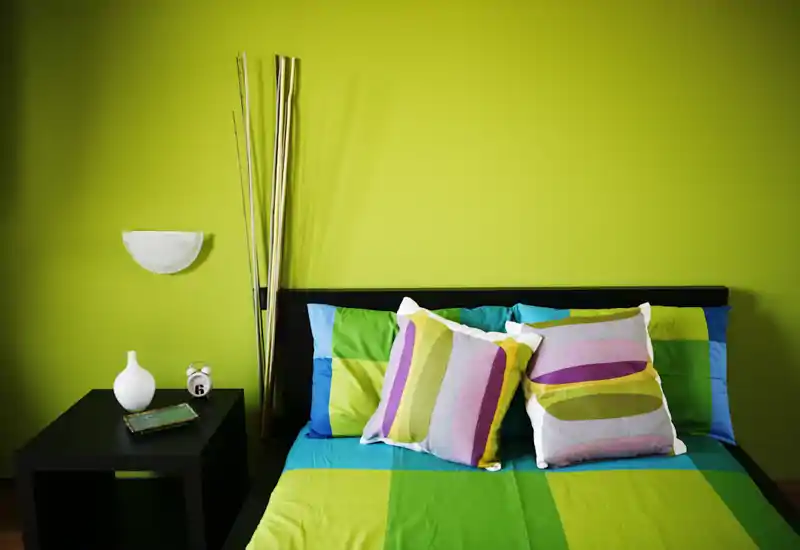 Camera da letto moderna e luminosa con un letto matrimoniale colorato contro una parete verde lime; cuscini a righe e biancheria coordinata donano un aspetto fresco e di design.