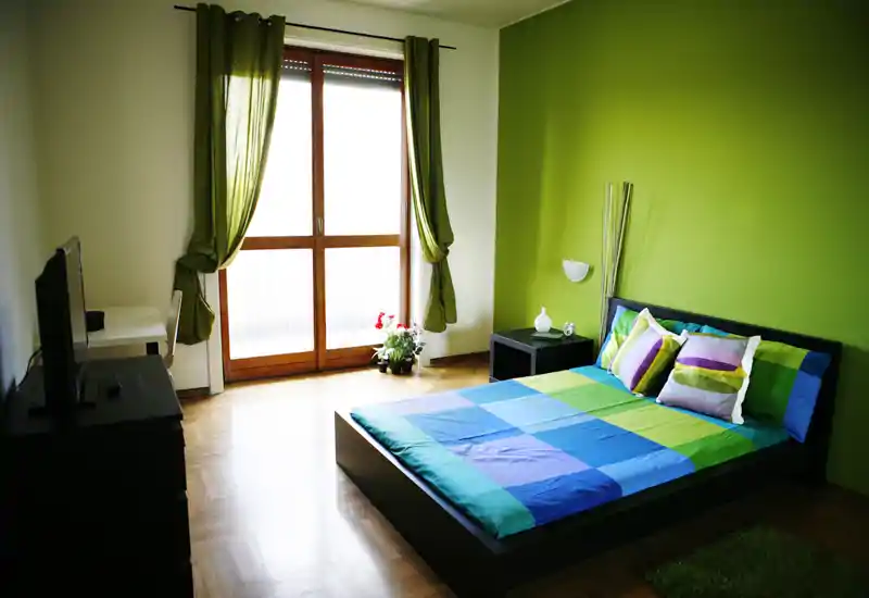 Camera da letto luminosa e moderna con letto matrimoniale, biancheria colorata e ampia finestra che lascia entrare molta luce naturale.