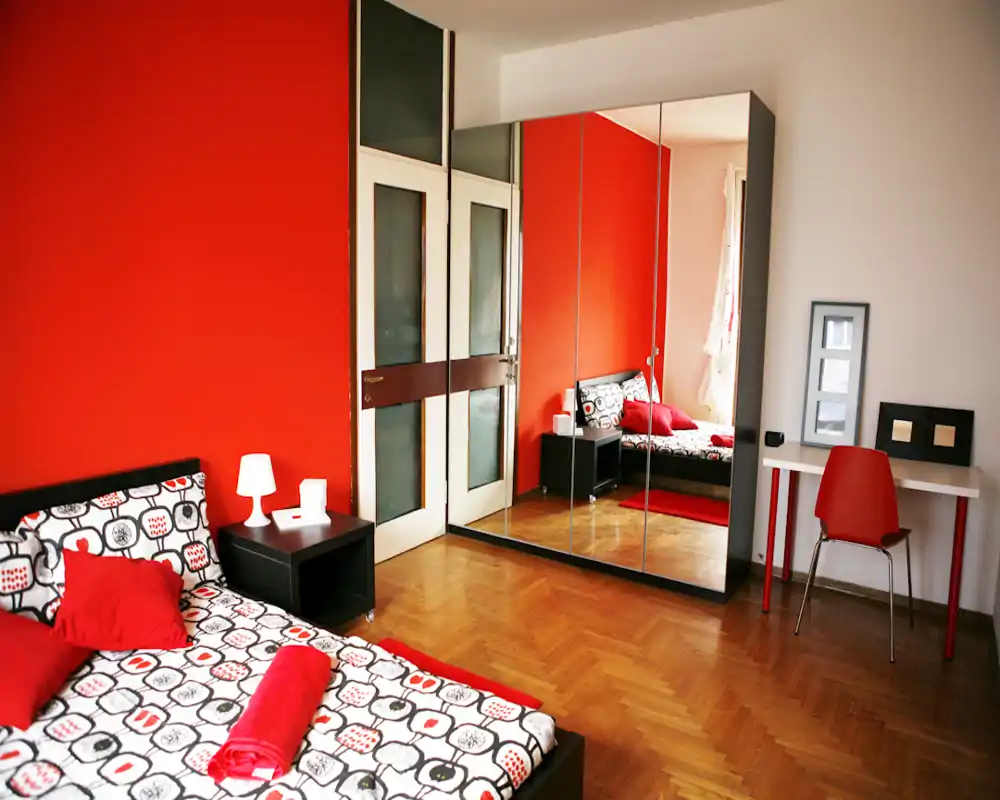 Chambre lumineuse et élégante avec lit double, linge de lit à motif et grande armoire miroir réfléchissant la pièce; mur rouge chaud et petit bureau pour une atmosphère cosy et moderne.