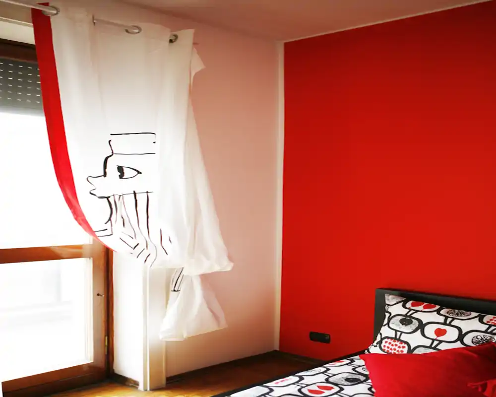 Coin chambre lumineux et moderne avec une couette à motifs, un mur d'accent rouge et une grande fenêtre habillée d'un rideau blanc laissant passer la lumière naturelle.