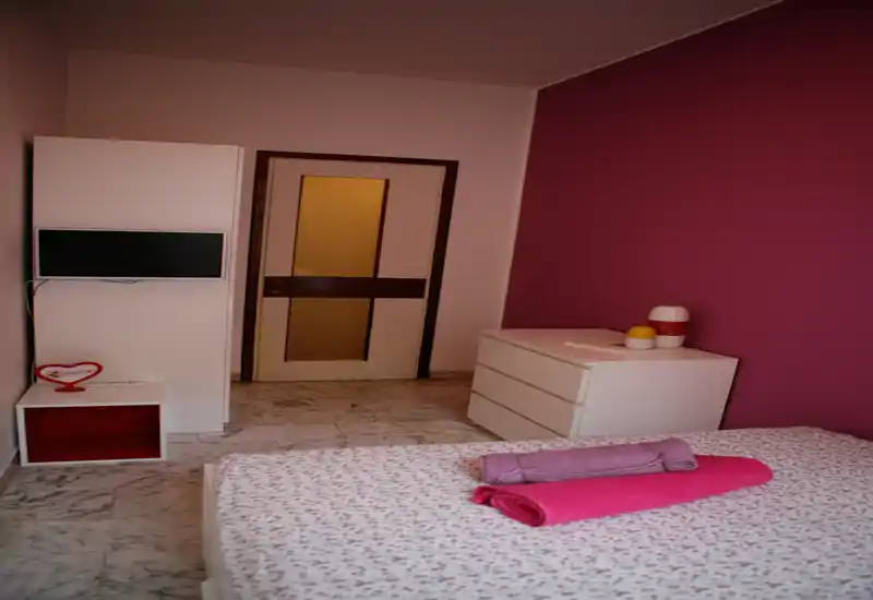 Bright, cozy bedroom with a patterned bedspread, neatly folded towels on the bed, a white dresser and contrasting pink accent wall — ideal for a welcoming rental listing.