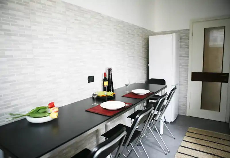 Angolo pranzo luminoso e moderno con un lungo bancone nero apparecchiato per due, sedie pieghevoli e frigorifero vicino — stile minimale e pulito, ideale per un monolocale.