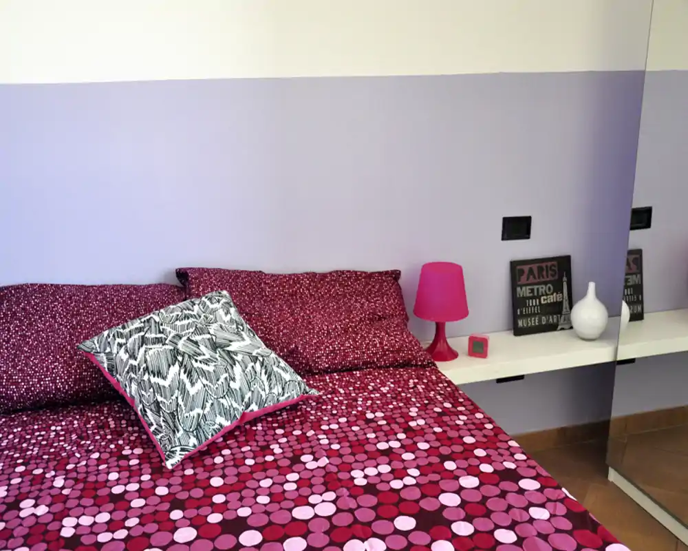 Camera da letto luminosa e alla moda con copriletto rosa a fantasia, cuscino decorativo e mensola con lampada e accessori.