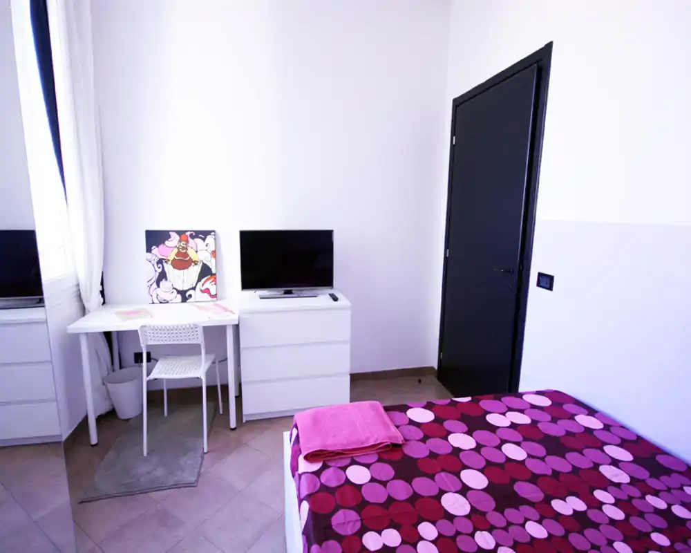 Camera da letto luminosa e moderna con letto matrimoniale a pattern, scrivania semplice e TV — biancheria fresca e luce naturale la rendono accogliente.