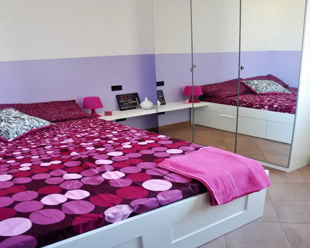 Camera da letto luminosa e moderna con letto contenitore bianco, biancheria rosa-viola a pois, plaid coordinato e lampada: l'armadio con ante a specchio amplia la stanza.