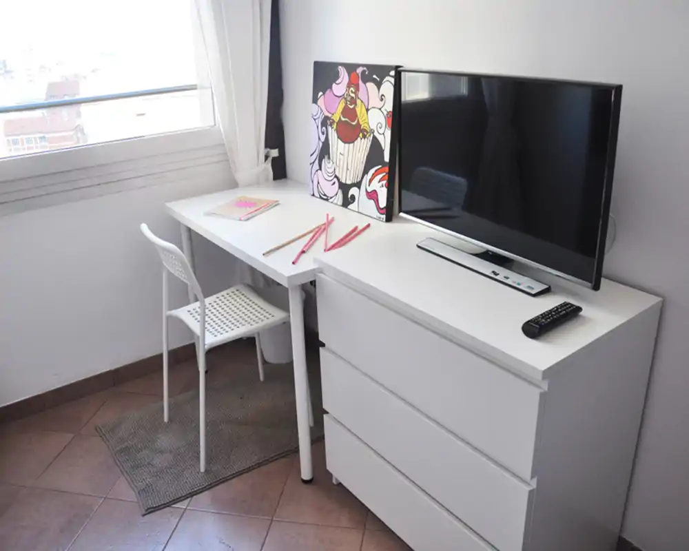 Angolo luminoso e minimal con scrivania bianca, sedia e comò basso con TV e un dipinto colorato — un angolo lavoro/soggiorno compatto e ben illuminato.
