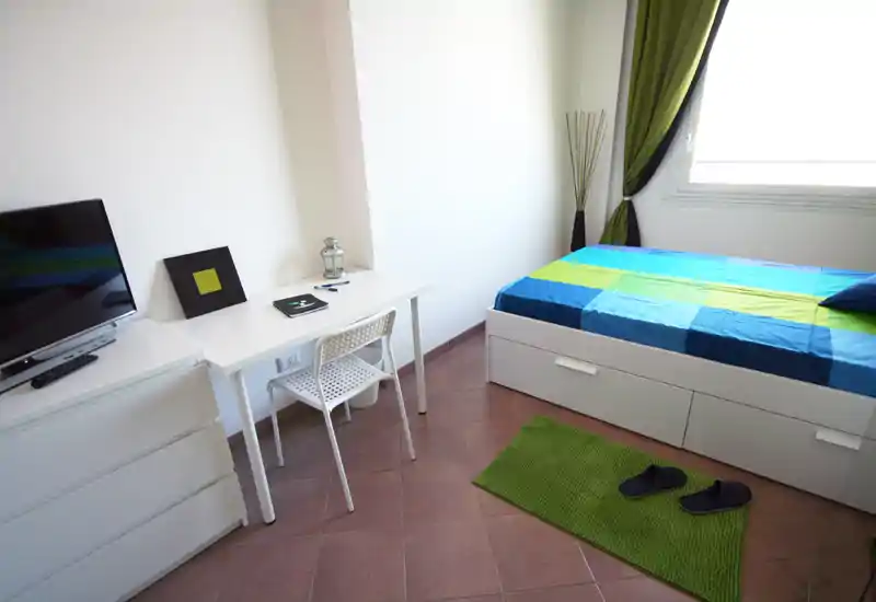 Dormitorio luminoso y moderno con cama individual con cajones, un pequeño escritorio y cómoda. Ropa de cama fresca, alfombra verde y luz natural desde la ventana crean un espacio limpio y acogedor, perfecto para un anuncio.