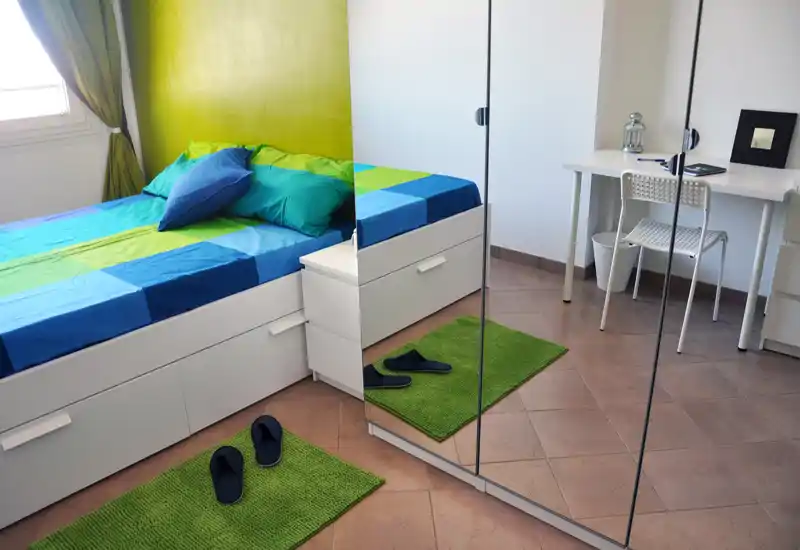 Dormitorio luminoso y moderno con ropa de cama verde y azul, cama con almacenamiento blanca, armario con espejo y un pequeño escritorio — ideal para una persona o huéspedes.